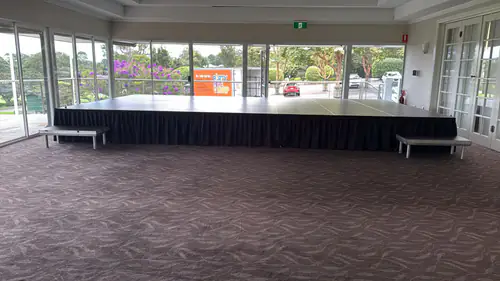 Large indoor stage platform with black skirting set up across a spacious function room with floor to ceiling windows
