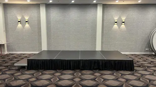 Low-rise modular event stage with black skirting set up in a function room with patterned carpet and wall lighting