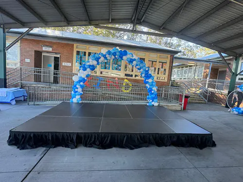 Outdoor event stage with black skirting set under a covered area, featuring a blue and white balloon arch backdrop at a school function