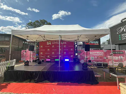 Outdoor event stage setup with performance platform, lighting effects, speakers, and branded backdrop under an open canopy structure