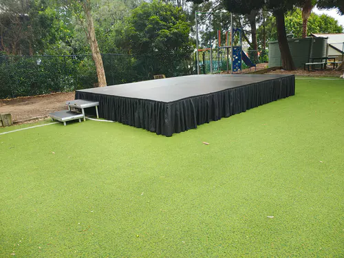 Outdoor stage platform with black skirting and access stairs positioned on artificial grass near a school playground area