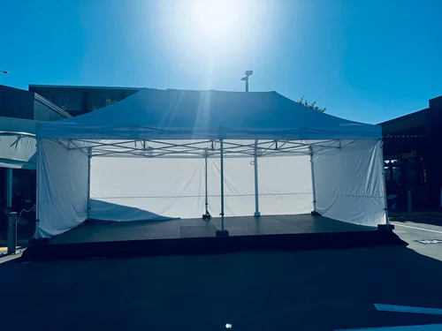 Outdoor stage platform positioned under a white marquee tent structure with side walls, set up for an event in a sunlit open area
