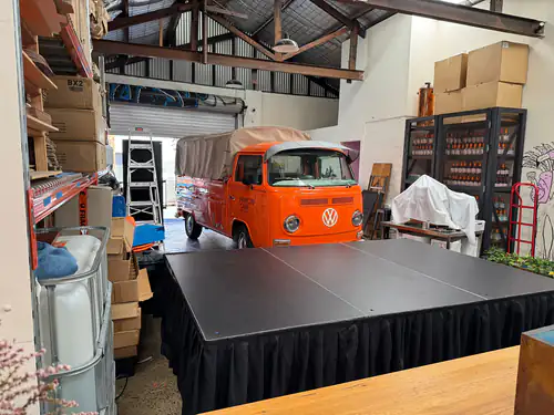 Portable stage platform with black skirting set up inside a warehouse space with equipment storage and display area in the background