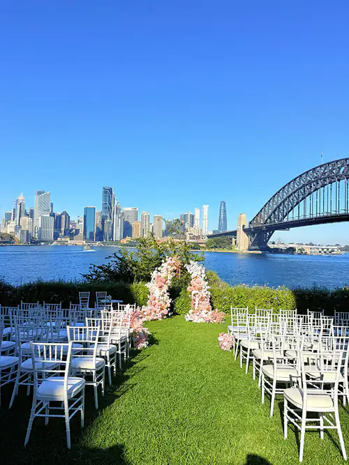 Lots of white Tiffany chairs set up for a wedding in a park in front of the Harbour Bridge