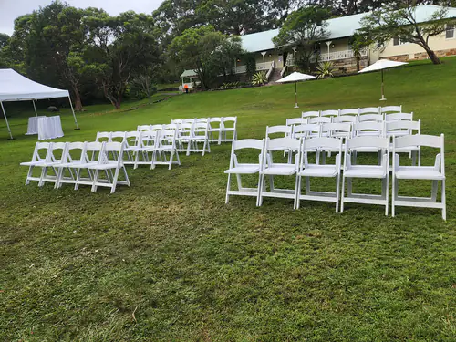 Scores of white folding plastic chairs in an outdoor park next to a marquee