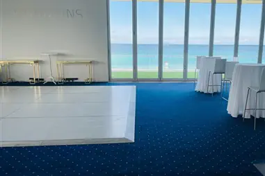 A white gloss dance floor positioned in a modern event space with floor-to-ceiling windows overlooking the ocean, featuring blue carpet and white cocktail tables with chairs