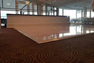 A white gloss dance floor positioned on brown patterned carpet with circular swirl designs in a modern venue space with large windows and decorative ceiling features