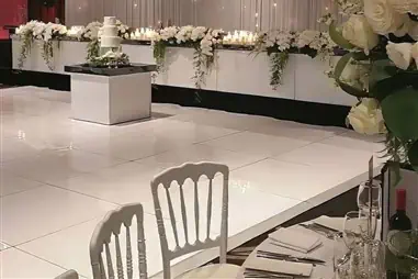 A pristine white gloss dance floor positioned in front of an elegant backdrop featuring cascading white florals, fairy light curtains, and white chiavari chairs in an upscale event venue