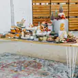 A White Hairpin Banquet Table with a Timber Top covered in a variety of delicious food, set on a rug in front of a brick wall.