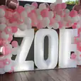 A pink and white balloon arch with the name Zoe illuminated by glow letters.