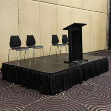 Black stage platform with a freestanding podium and four black chairs set up in an indoor event room with patterned carpet and neutral wall panels