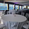 A room with white tables and chairs, adorned with white round banquet tablecloth hire, offering a stunning view of the ocean.