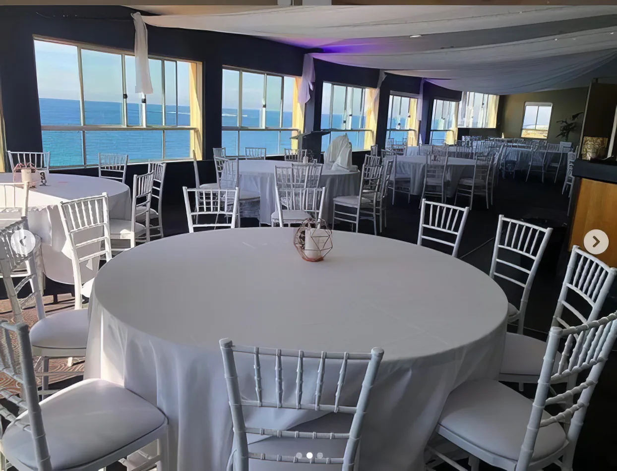 A room with white tables and chairs, adorned with white round banquet tablecloth hire, offering a stunning view of the ocean.
