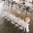 A white table with white chairs adorned with greenery hanging from the ceiling.