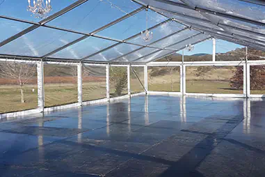 Clear roof event marquee with transparent walls, black flooring, exposed frame, chandelier lighting, and open views across a rural outdoor setting