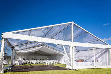 Clear roof framed marquee with enclosed transparent side walls, draped ceiling lining, dining tables and timber flooring, providing all-weather shelter while maintaining natural light and open views