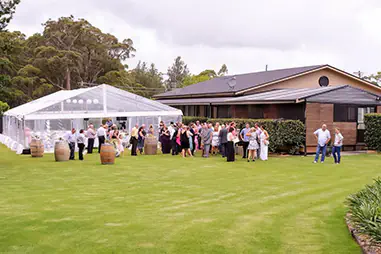 Clear wall framed marquee set up beside a lawn with wedding guests gathered outside, timber barrel decor and white event seating visible inside