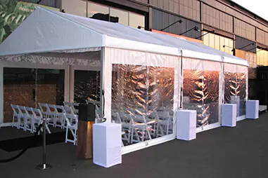 Framed marquee exterior with clear side walls, white roof panels, white cube seating, rows of chairs inside and a roped entry area beside an event venue