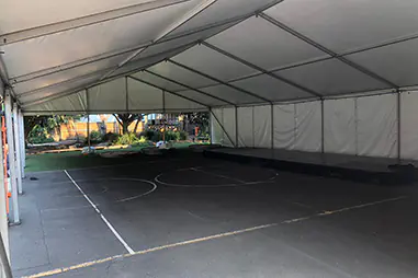 Large school event marquee set up over an outdoor court with white roof panels, solid side walls, open end access and a low black stage platform
