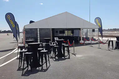 Marquee hire setup for an airport event with a large white structure, clear side panels, black cocktail tables, bar stools, flags, and roped entry area