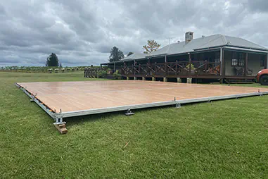 Large outdoor event flooring platform installed on grass beside a rural venue, with timber-look panels, metal edging, and a surrounding vineyard landscape