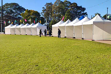 Row of white event stall marquees with side walls set up along a grass area, providing covered weather protection for outdoor vendors, registration points or service booths