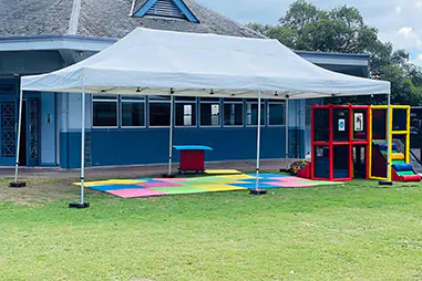 White school event canopy set up outside a school building with colourful soft play mats, play equipment and open lawn space for a children’s activity area