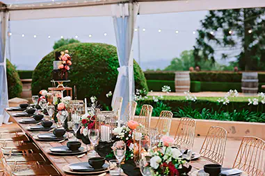 Wedding marquee dining setup with a long banquet table, gold wire chairs, black tableware, floral centrepieces, candle lanterns, clear side walls and festoon lighting overlooking landscaped gardens