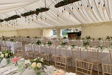 Wedding marquee hire setup with long banquet tables, white tablecloths, timber chairs, floral centrepieces, draped ceiling lining, hanging pendant lights and greenery-covered timber beams