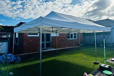 White backyard gazebo canopy set up over a lawn beside a brick house, with weighted legs and open sides for outdoor shade coverage