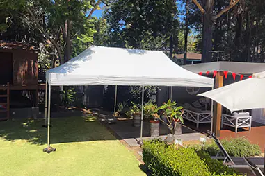 White backyard pop-up canopy set up over a garden lawn beside an outdoor seating area, providing shade for a casual private event