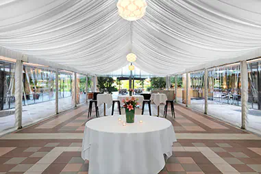 White draped marquee interior with clear side walls, round cocktail tables, black stools, patterned floor tiles and suspended pendant lighting