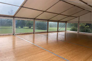 White event marquee interior with timber flooring, clear window walls, white roof panels, and open lawn views outside