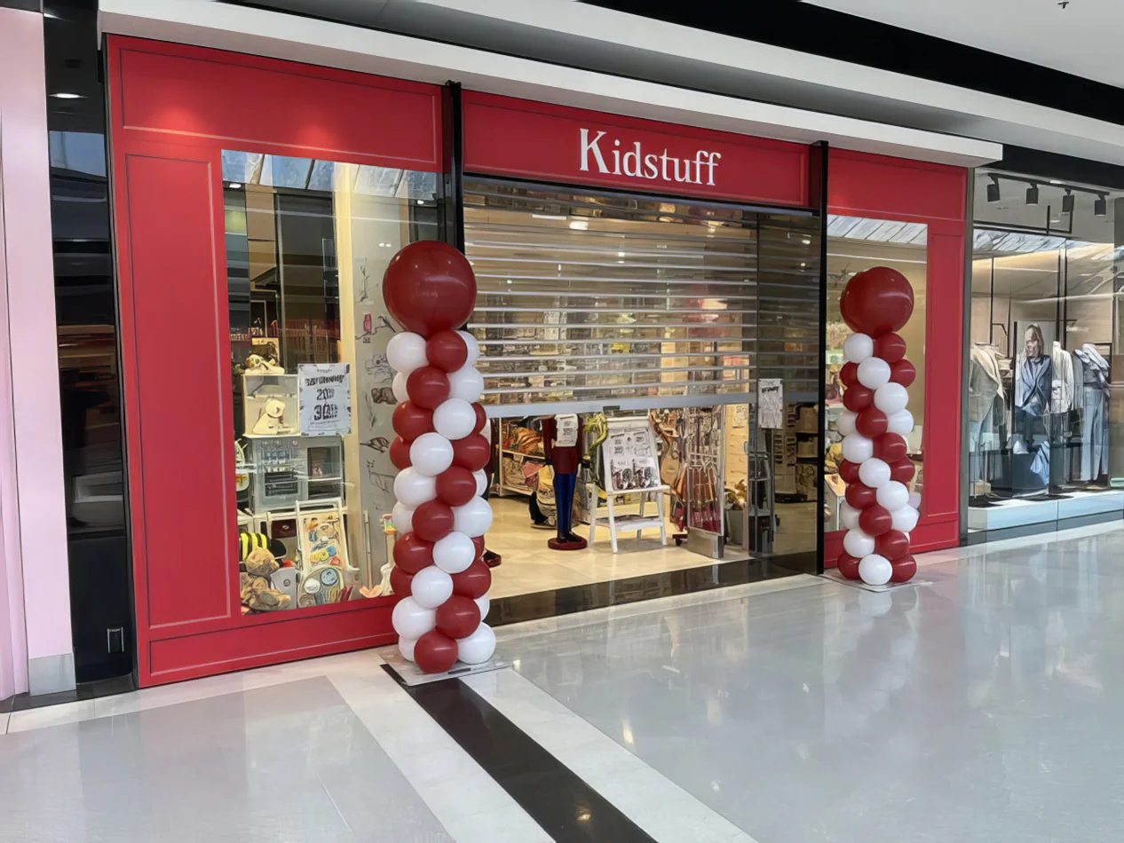 Red and white spiral balloon pillars placed on either side of a toy store entrance in a shopping centre, adding a festive decorative display suitable for retail promotions or event theming.