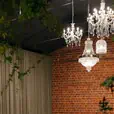 Natural timber bentwood chairs arranged along white linen-covered banquet tables in an elegant event space, with crystal chandeliers overhead, exposed red brick walls, draped sage curtains, and trailing greenery
