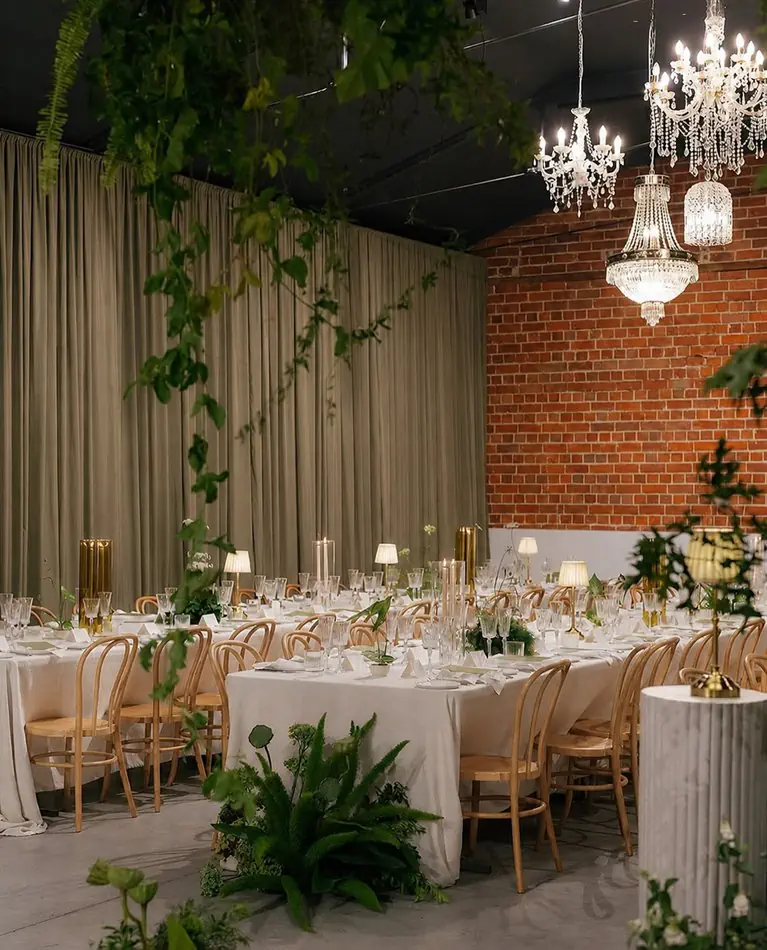 Natural timber bentwood chairs arranged along white linen-covered banquet tables in an elegant event space, with crystal chandeliers overhead, exposed red brick walls, draped sage curtains, and trailing greenery
