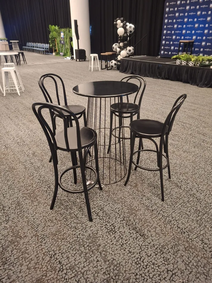 Four black bentwood bar stools with curved back rests arranged around a black wire cocktail table at an indoor sports event