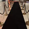 A long black carpet runner stretching through an indoor venue, flanked by gold stanchion posts connected with black velvet ropes, with a gathering of people and warm amber lighting visible in the background
