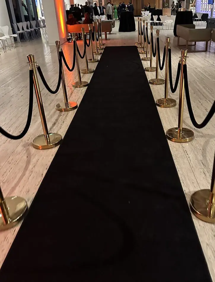 A long black carpet runner stretching through an indoor venue, flanked by gold stanchion posts connected with black velvet ropes, with a gathering of people and warm amber lighting visible in the background