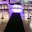 A black carpet runner leads into an aviation hangar dressed for a Lamborghini event, flanked by rope and post barriers, with yellow and purple sports cars on either side and a private jet visible in the background beneath warm purple and amber lighting