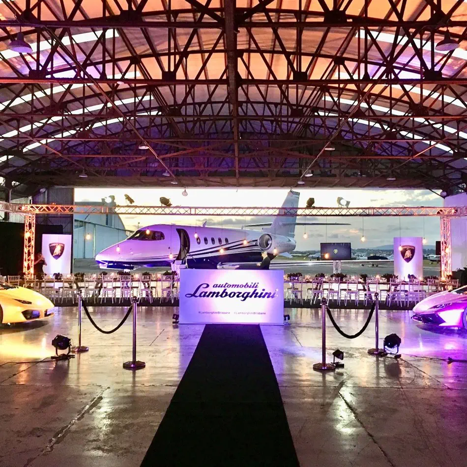 A black carpet runner leads into an aviation hangar dressed for a Lamborghini event, flanked by rope and post barriers, with yellow and purple sports cars on either side and a private jet visible in the background beneath warm purple and amber lighting