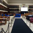 A black carpet runner laid down the centre of a grand library hall, flanked by silver stanchions with rope barriers, blue event furniture, and towering wooden bookshelves lining the walls