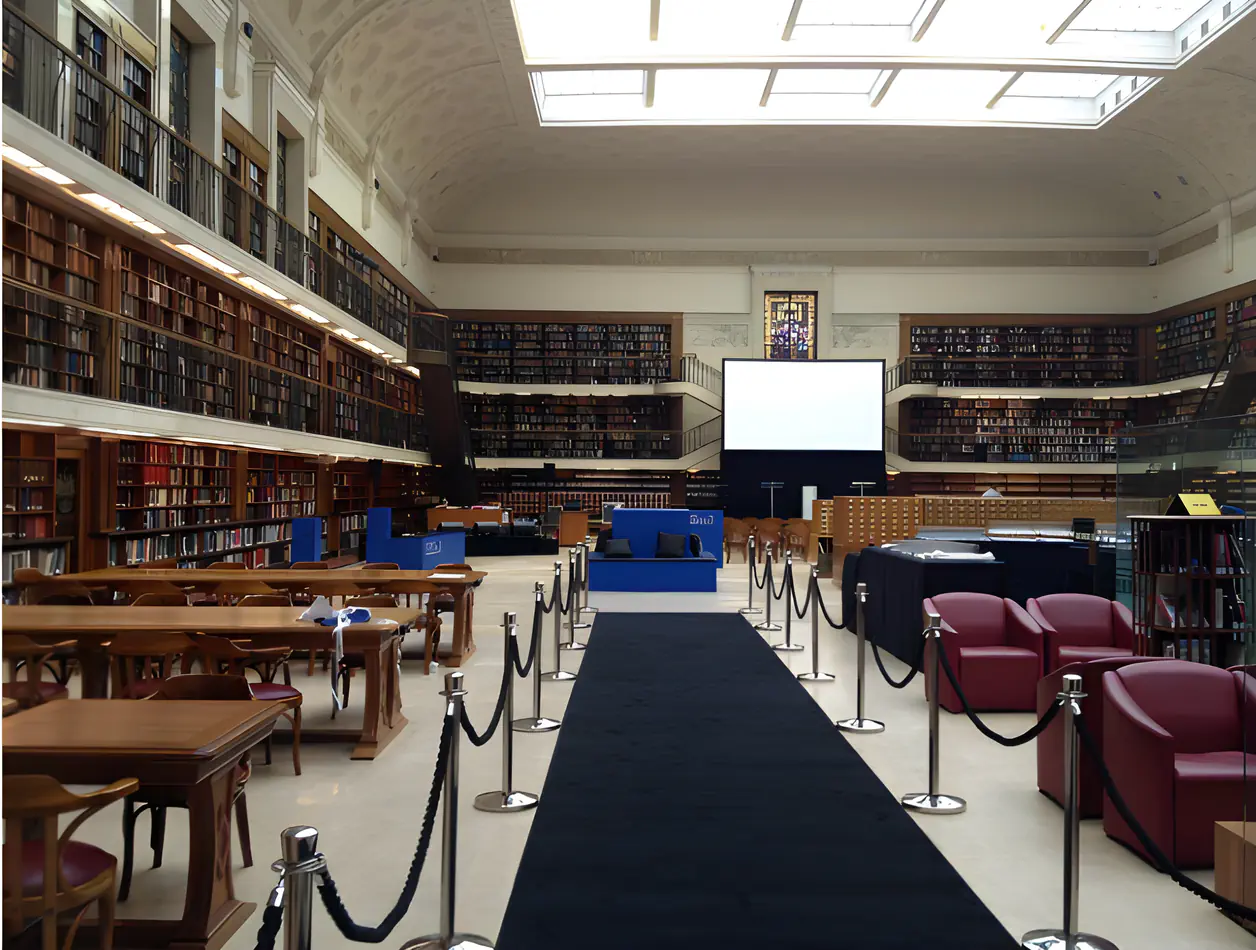 A black carpet runner laid down the centre of a grand library hall, flanked by silver stanchions with rope barriers, blue event furniture, and towering wooden bookshelves lining the walls