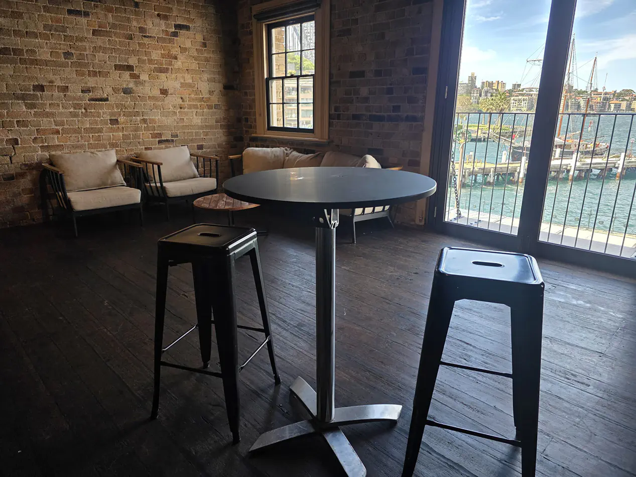 Black cocktail table with industrial black bar stools positioned inside a harbour-side function venue with exposed brick walls