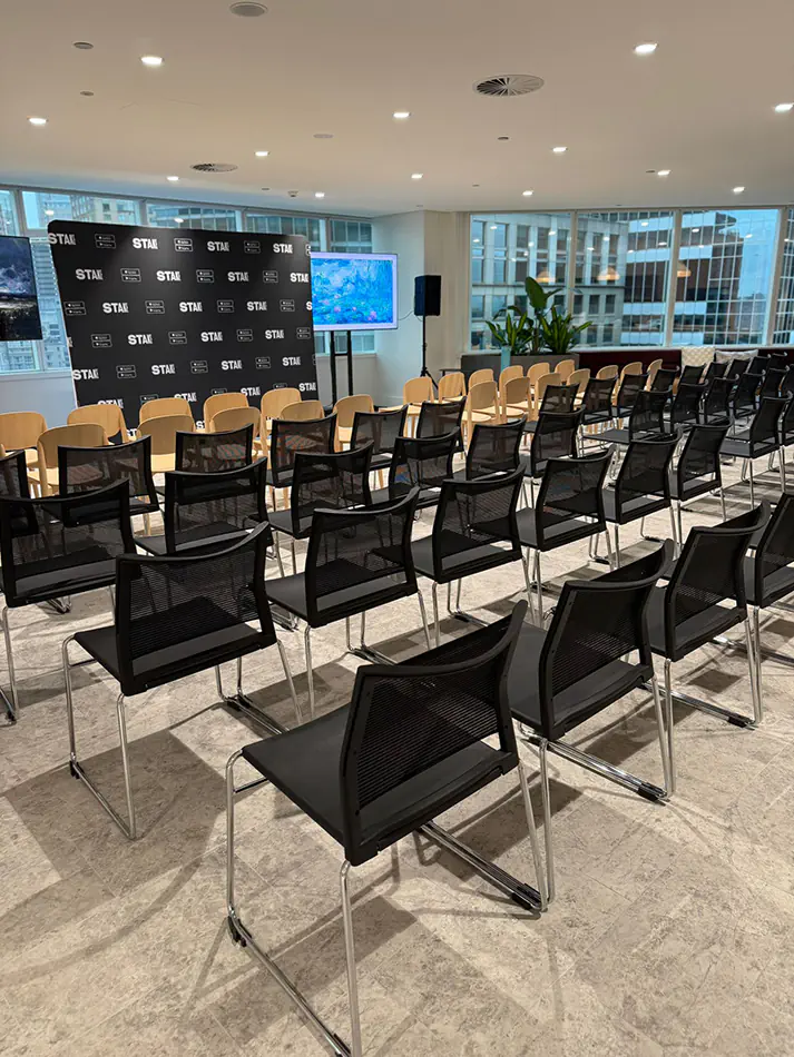 Black conference chairs arranged for a corporate presentation event with stage backdrop, display screens and city office views