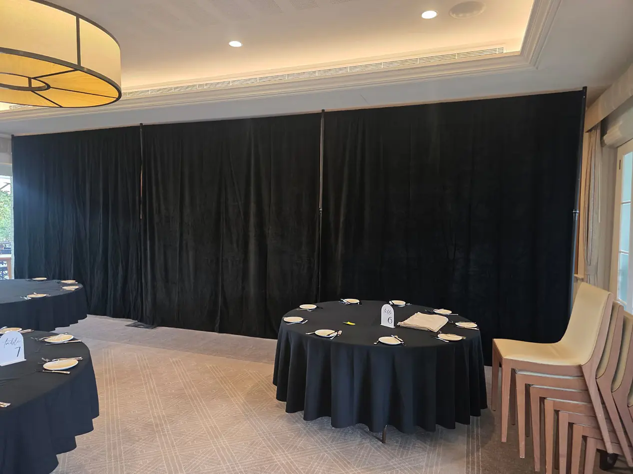 Black event draping across a banquet room wall with round dining tables, black tablecloths and neutral carpet flooring