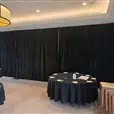 Black event draping across a banquet room wall with round dining tables, black tablecloths and neutral carpet flooring