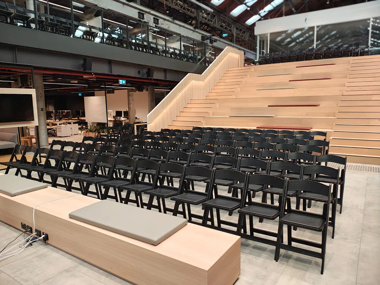 Rows of black folding chairs with padded seats arranged for event seating inside a modern venue with tiered timber steps and open interior space