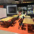 Event setup featuring multiple black hairpin banquet tables with natural timber tops, paired with black folding chairs on an orange floor covering, arranged in a retail venue space for a workshop or gathering.