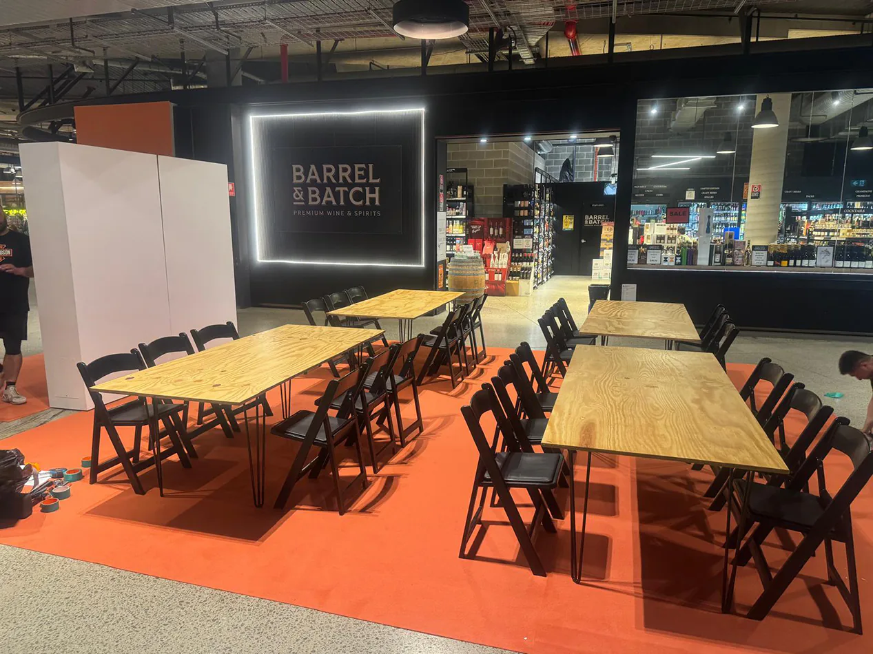 Event setup featuring multiple black hairpin banquet tables with natural timber tops, paired with black folding chairs on an orange floor covering, arranged in a retail venue space for a workshop or gathering.